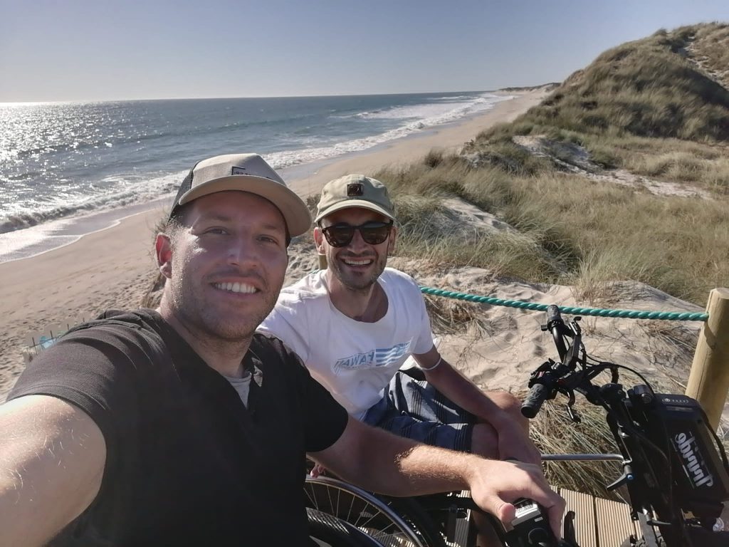 Zwei Männer mit Basecap lachen in die Kamera. Sie sitzen im Rollstuhl mit Elektromotor. Hinter ihnen sieht man das Meer und den Sandstrand. Die Sonne scheint.