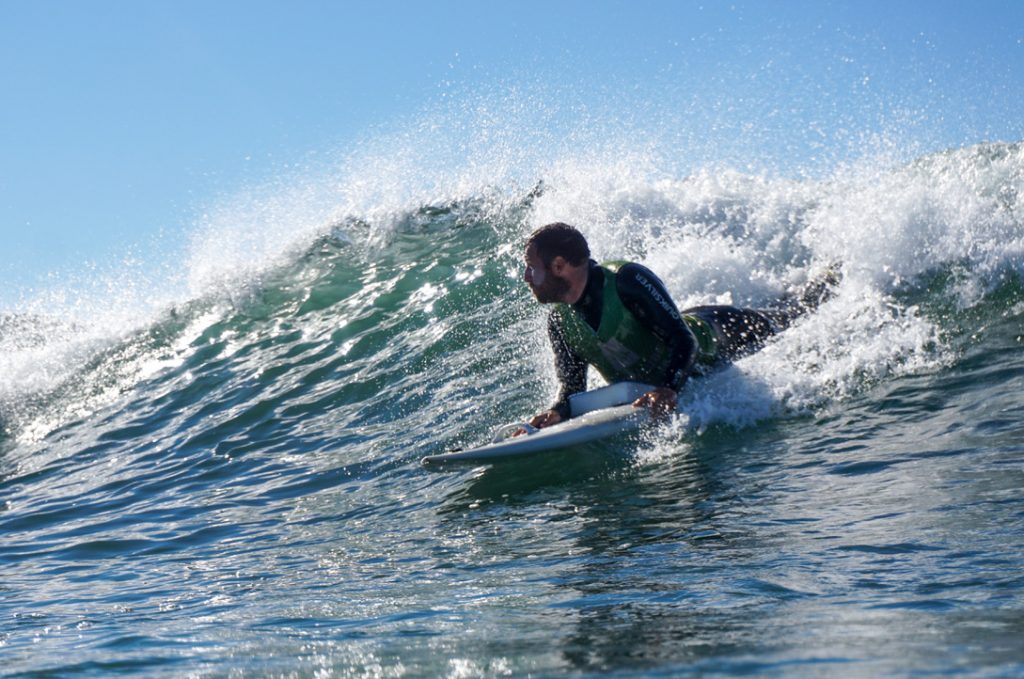 Ein Mann surft auf dem Bauch liegend eine grüne Welle. Hinter ihm spritz das Weißwasser hoch. Sein Blick ist nach rechts gerichtet und er drückt seinen Oberkörper weit nach oben, während er sich mit beiden Händen rechts und links am Brett festhält. Unter seiner Brust sieht man einen Schaumstoffkeil, der auf dem Brett befestigt ist.