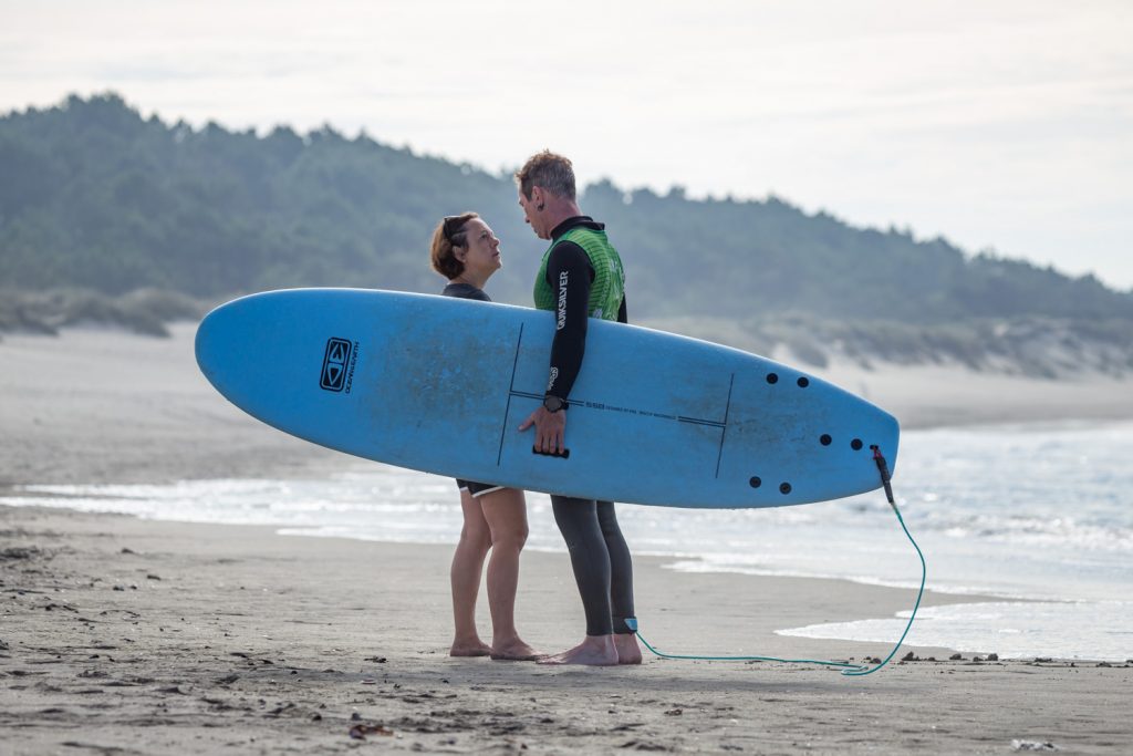 Eine Frau und ein Mann stehen sich am Strand gegenüber und blicken einander in die Augen. Der Mann hat ein blaues Surdboard under dem Arm.
