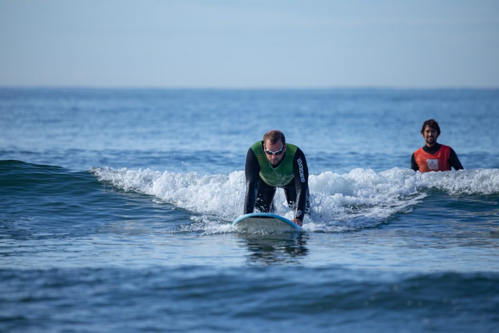 Ein Mann mit Schwimmbrille kniet auf dem Surfbrett und surft eine Weißwasserwelle. Im Hintergrund sieht man seinen Surfcoach, der lächelt.