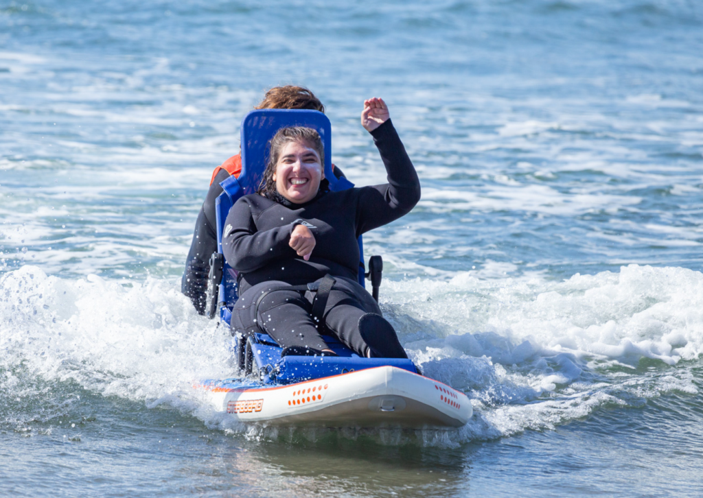 Eine Frau im Neoprennzug sitzt auf einem Surfbrett im Meer. Sie hebt einen Arm in die Luft und lacht in die Kamera. Hinter ihr sieht man die Rückenlehne eines Sitzes und den Oberkörper einer Person, die sie durchs Wasser schiebt.