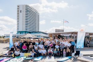 Einew große Gruppe von Menschen - manche stehen, andere sitzen im Rollstuhl. Vor ihnen liegen verschiedene Surfbretter.