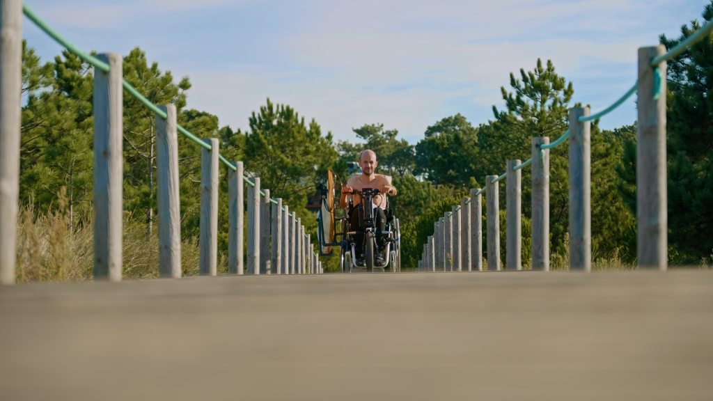Mann im Rollstuhl rollt auf Holzsteg entlang. An der Seite klemmt ein Surfbrett.