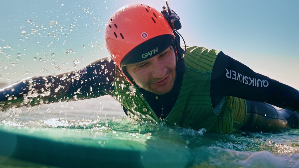 Person mit Helm und Headset in Nahaufnahme. Sie liegt auf einem Surfbrett und paddelt.