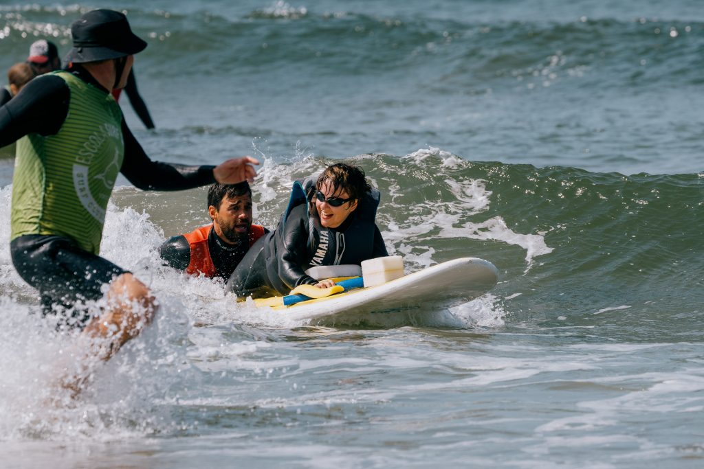 Eine Frau liegt bäuchlings auf einen Surfbrett und surft eine Welle. Auf dem Brett sieht man verschiedene Keile, die Frau trägt eine Schwimmweste, Schwimmbrille und lacht. Hinter ihr hält ein Mann das Surfbrett fest und surft mit ihr. Neben ihnen rennt eine weitere Person im Wasser.
