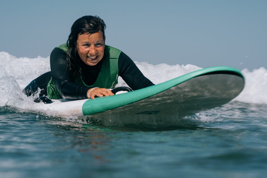 Eine Frau in Nahaufnahme. Sie liegt bäuchlings auf einen Surfbrett und surft eine Welle. Sie lacht.