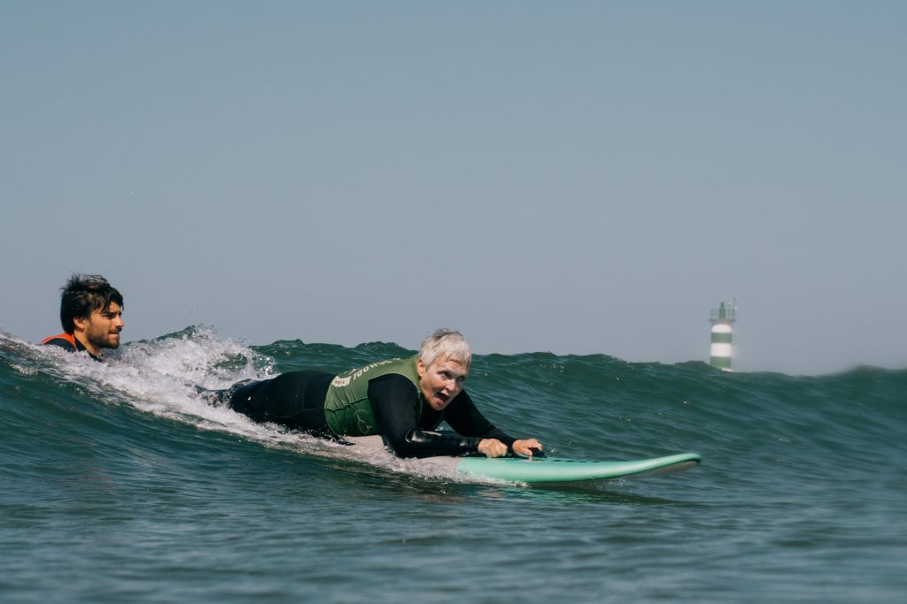 Eine Frau surft - auf dem Bauch liegend - eine Welle. Ihr Mund ist vor Erstauen geöffnet. Hinter ihr sieht man den Kopf eines Mannes, der ihr einen Schubs gibt.