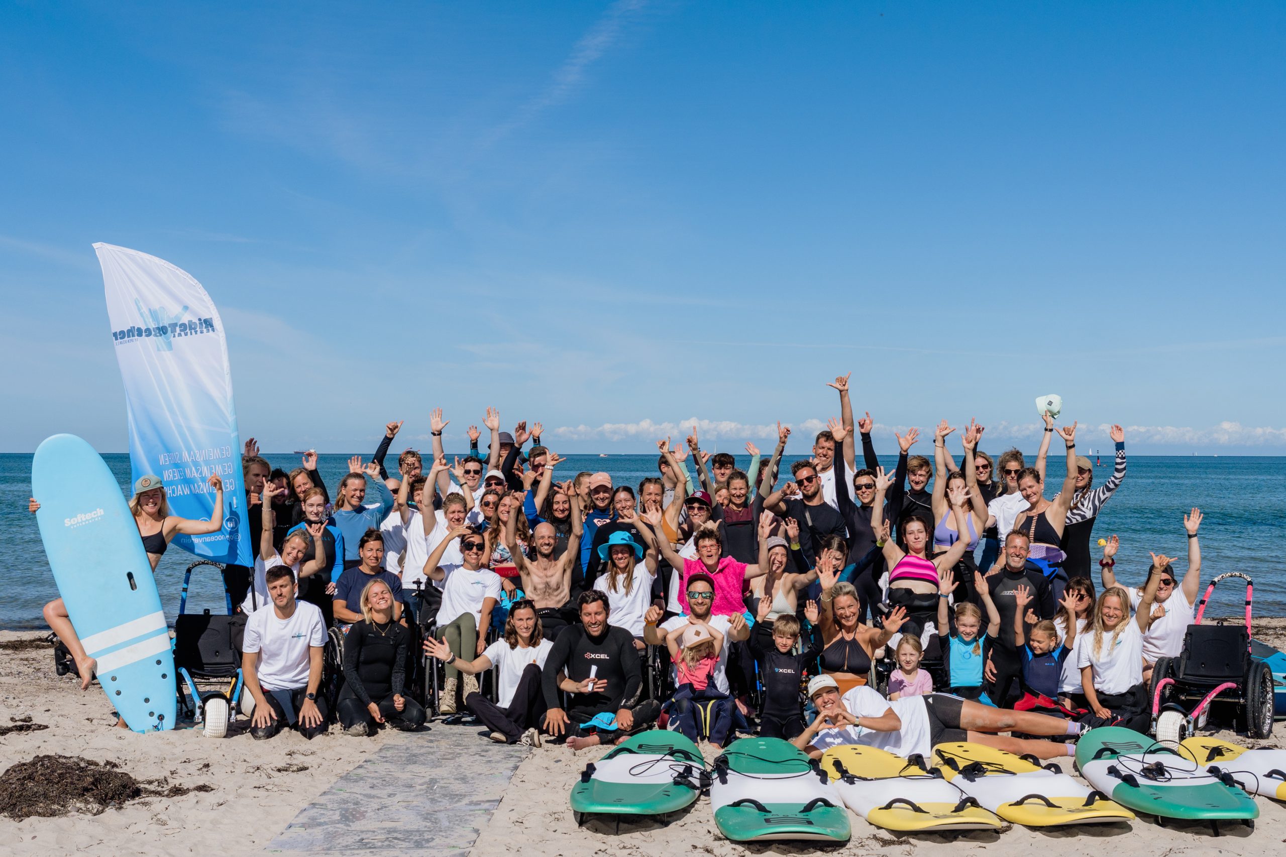 Eine große Gruppe von Menschen am Strand - hinter ihnen das Meer und blauer Himmel. Manche sitzen oder knien, andere stehen oder sitzen im Rollstuhl. Die Menschen reißen die Arme nach oben. Vor ihnen siehr man einige Surfbretter mit Haltegriffen und Beach-Flaggen.