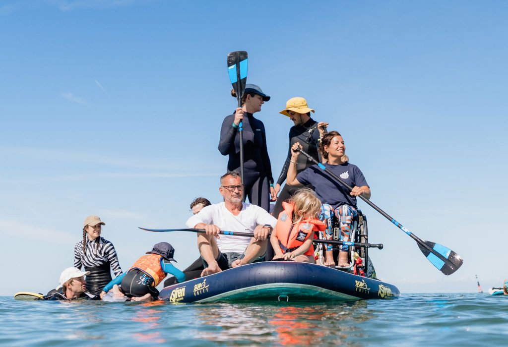 Eine Gruppe von Menschen - Erwachsene und Kinder - auf einem großen Stand-up-Paddleboard im Wasser. Einige stehen und haben Paddel in der Hand, andere sitzen. Eine Frau sitzt in ihrem Rollstuhl und paddelt. Links klettert ein Kind auf das Brett.